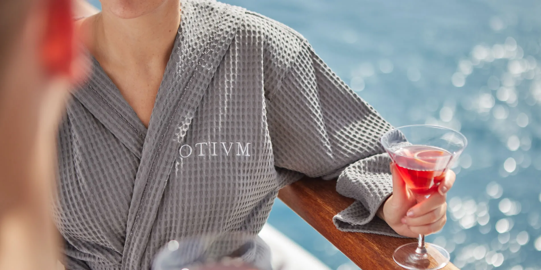 Person in gray Otivm bathrobe holding red cocktail by water