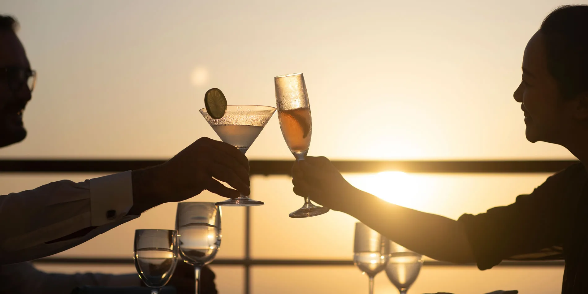 Two hands toasting cocktails at sunset with silhouettes