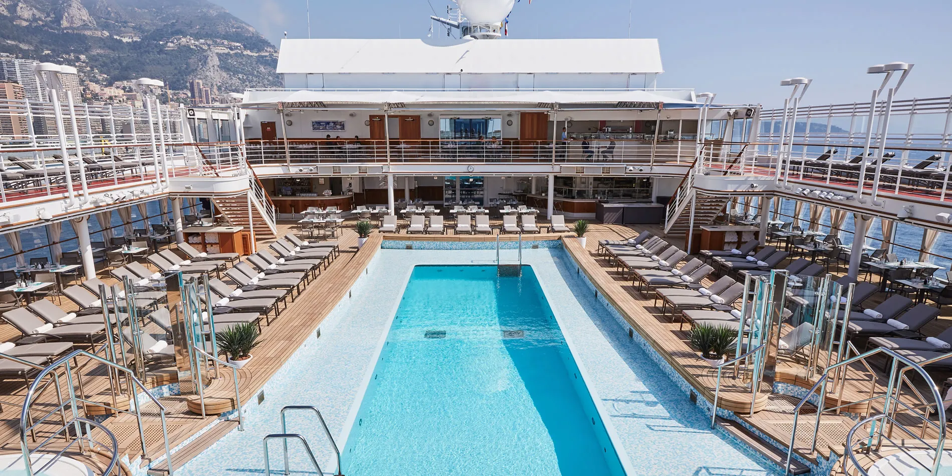Luxury cruise ship pool deck with lounge chairs overlooking mountainous coastline