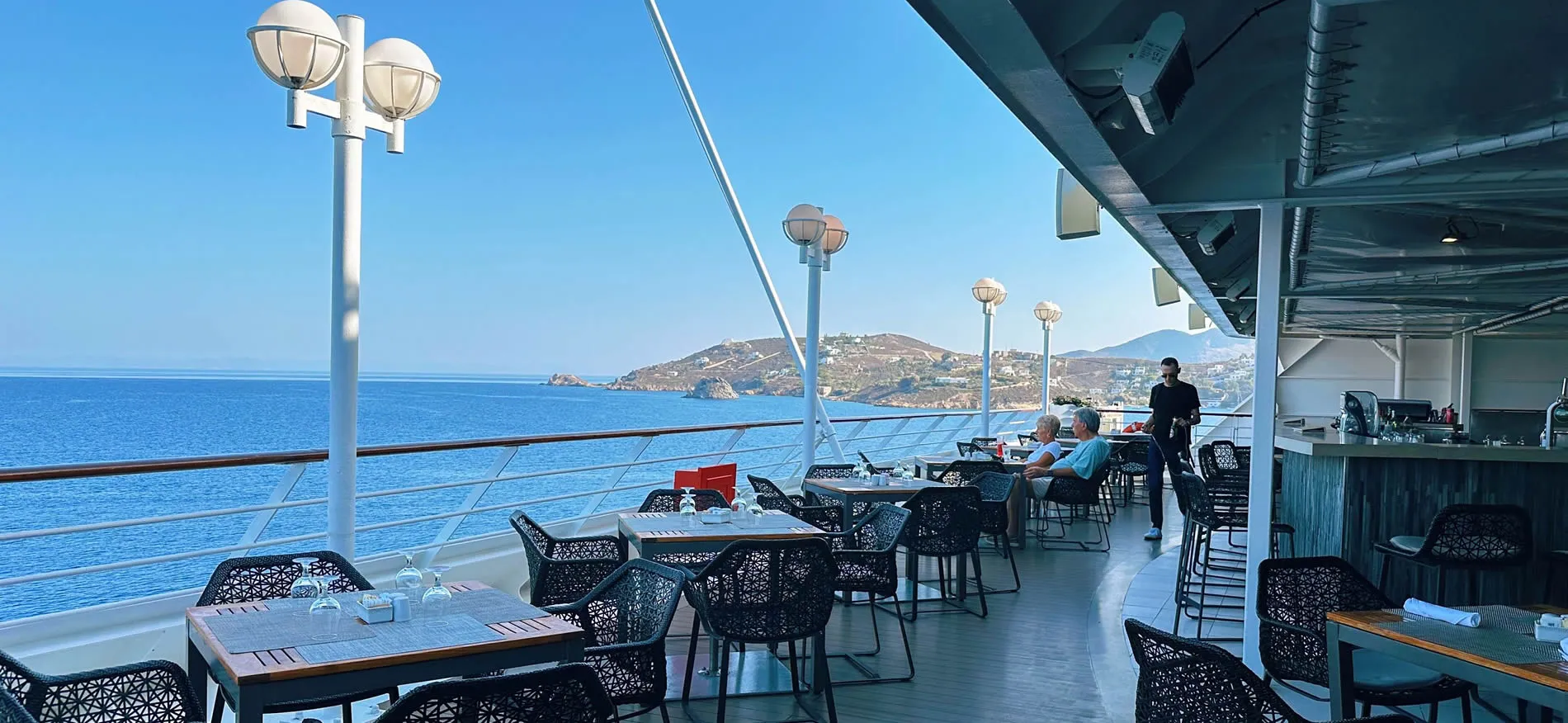 Cruise ship deck with ocean view, tables, and coastal landscape