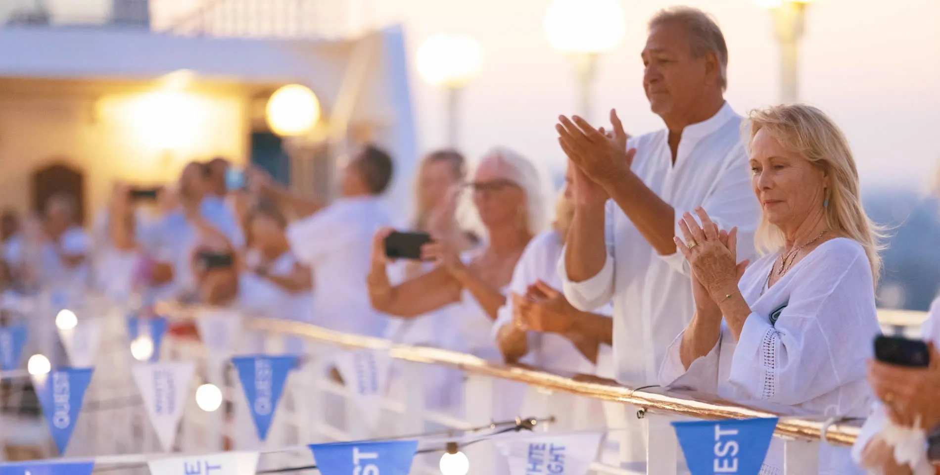 Crowd in white attire clapping at outdoor event with blue flags