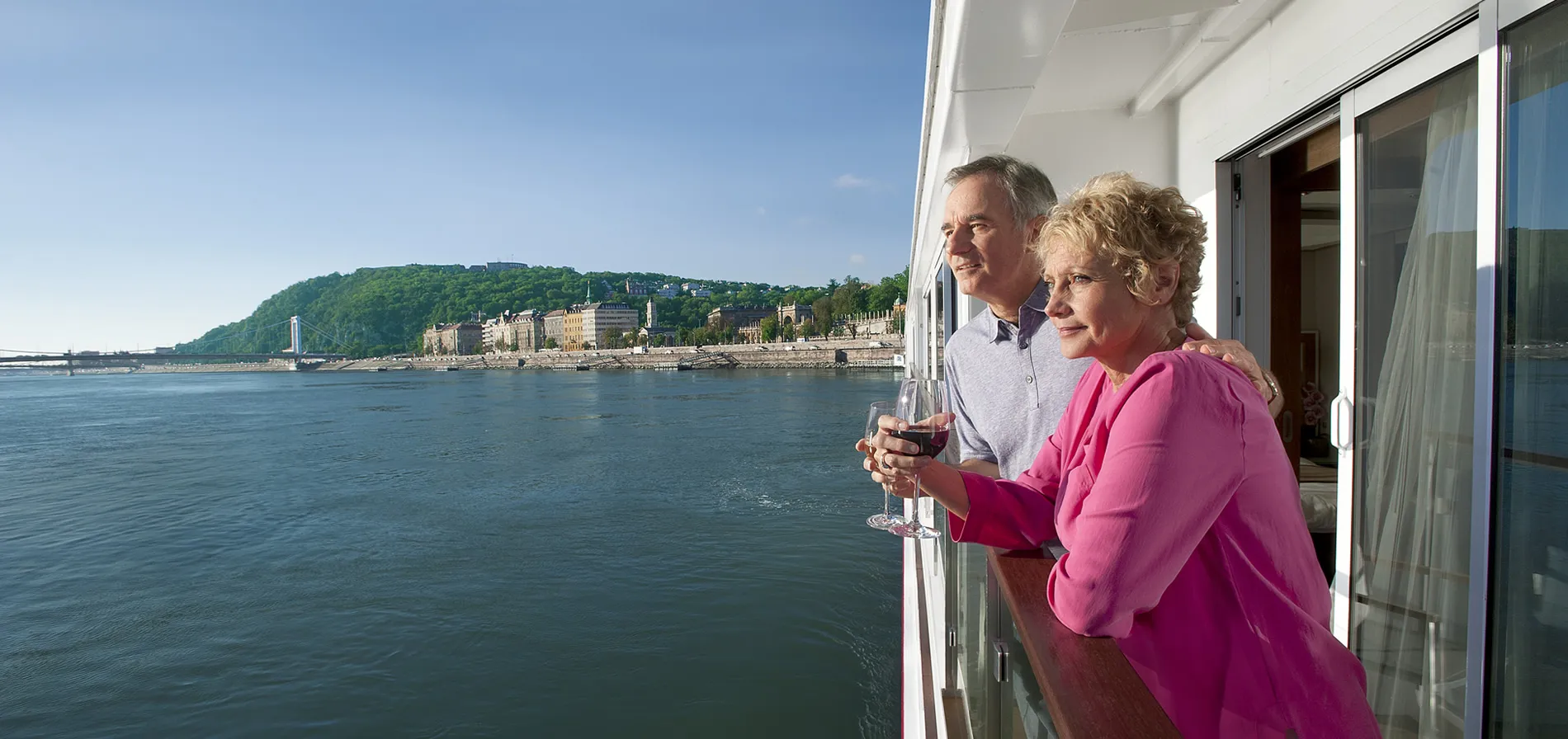 Elderly couple enjoying wine on cruise ship balcony overlooking riverside city