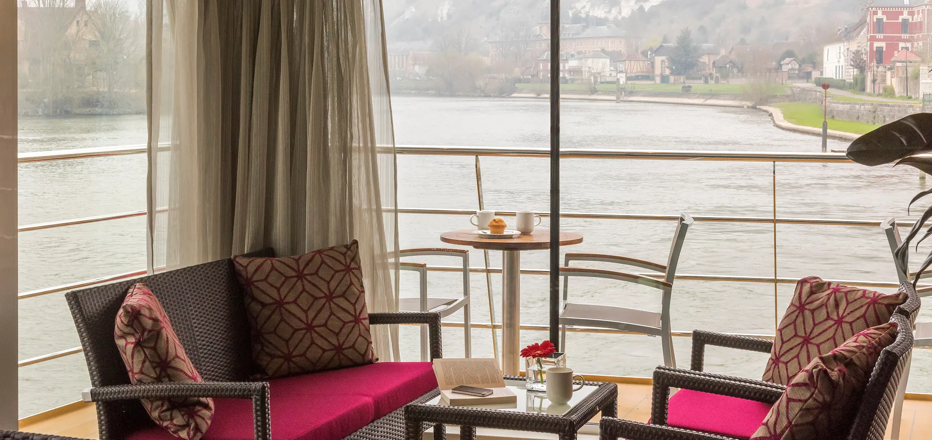 Cozy riverside seating area with coffee and pastry, viewed through sheer curtains