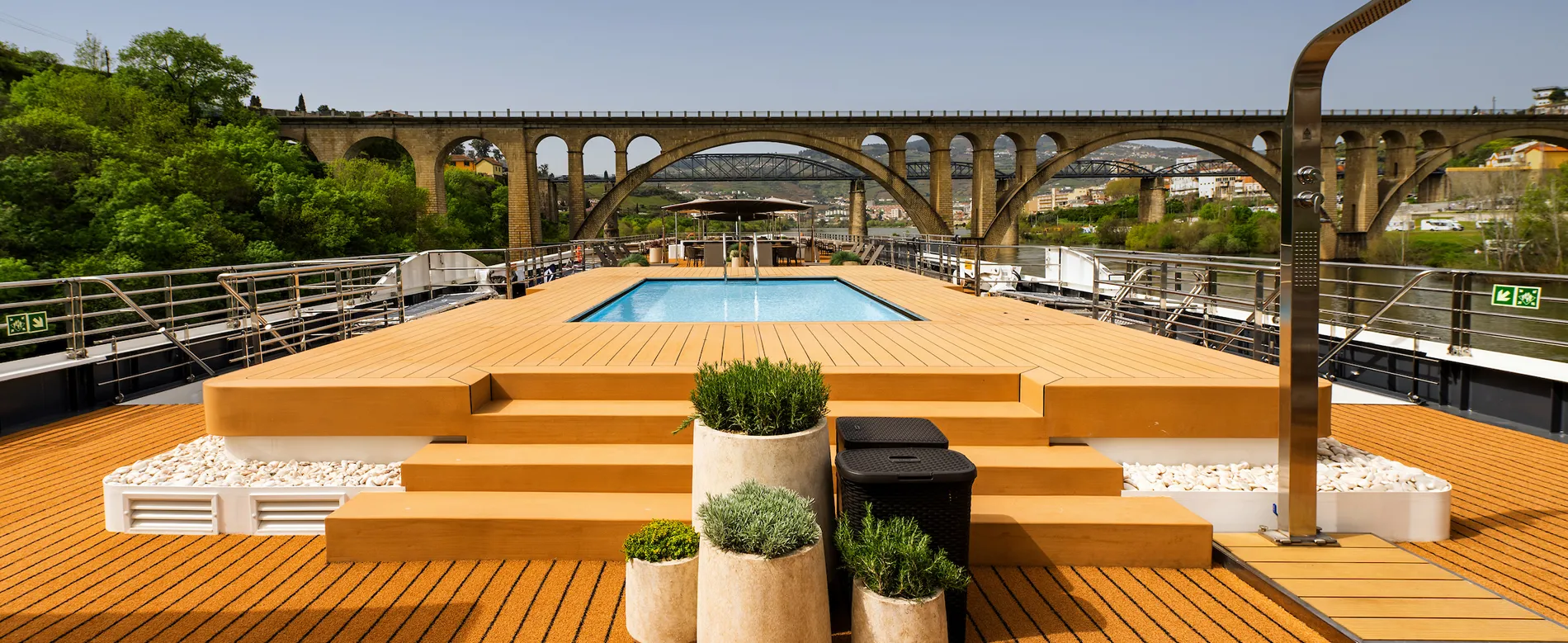 Modern pool deck with wooden platforms and stone planters, stone bridge background