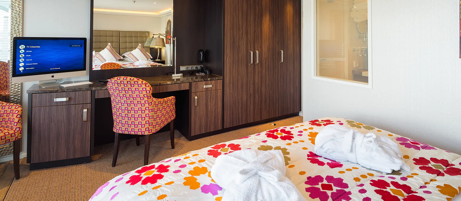 Colorful hotel room with floral bedspread, desk, TV, and folded white towels