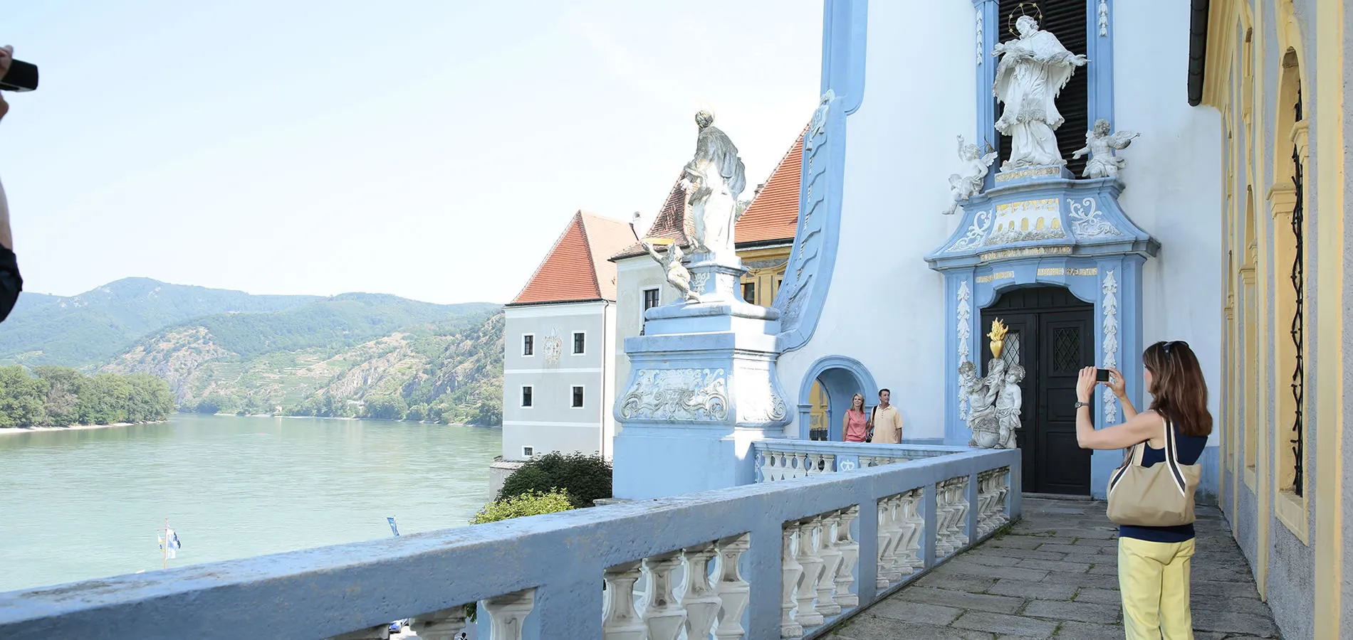 Blue baroque building with statues overlooking scenic river and mountain landscape