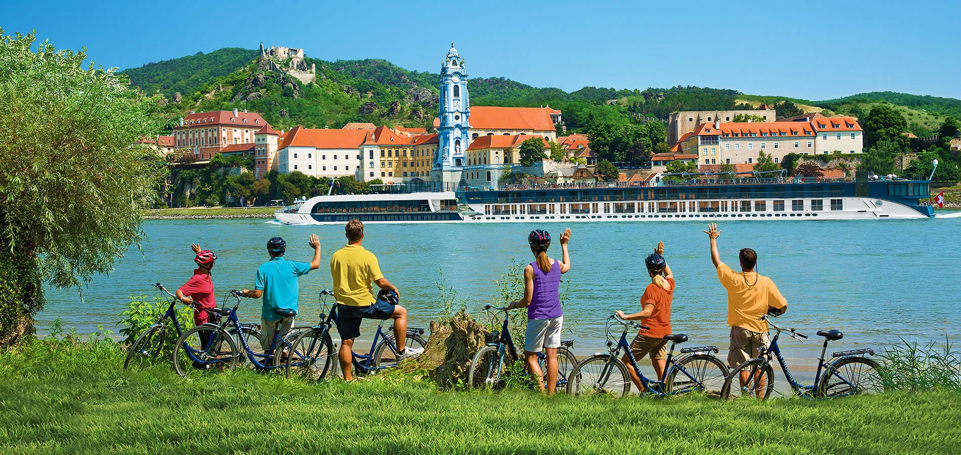 Cyclists wave to river cruise boat passing scenic European town with blue church