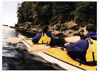 Two kayakers paddle along rocky shoreline with dense forest background