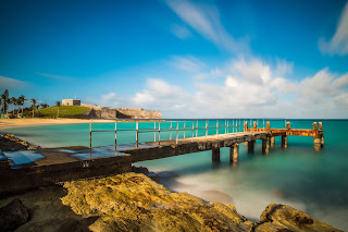 Wooden pier extending into turquoise ocean with rocky shoreline and blue sky