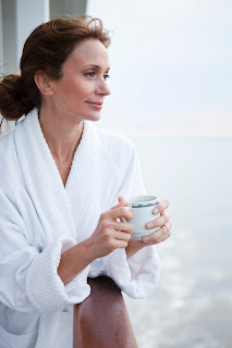 Woman in white bathrobe holding coffee mug, looking contemplative by window