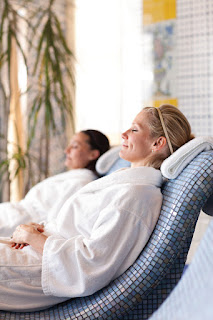 Two people in white robes relaxing at a spa, leaning back with closed eyes
