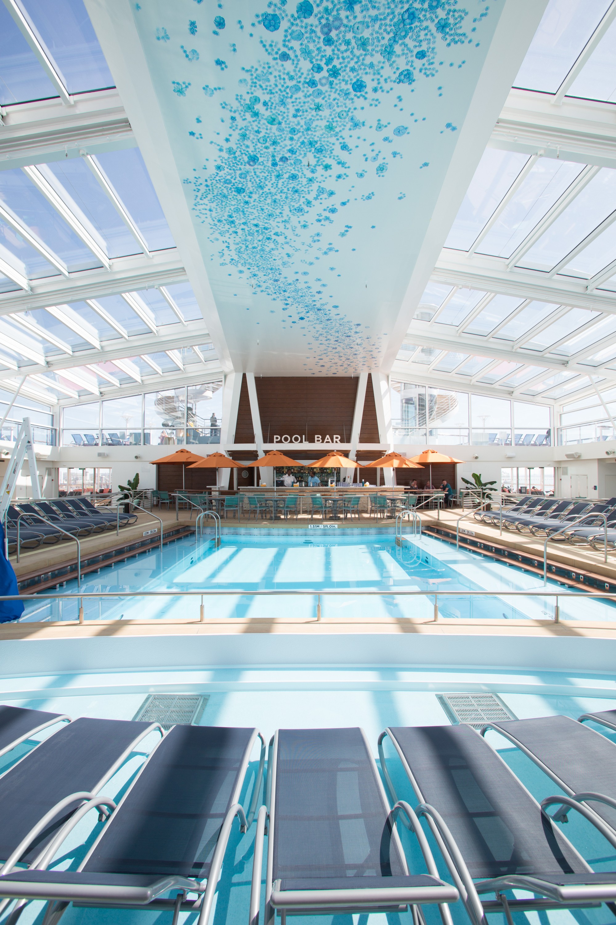 A wide editorial travel photograph of a busy cruise ship pool deck filled with families and guests of varying ages enjoying the sun, capturing the energetic and social atmosphere of a large ship at fu