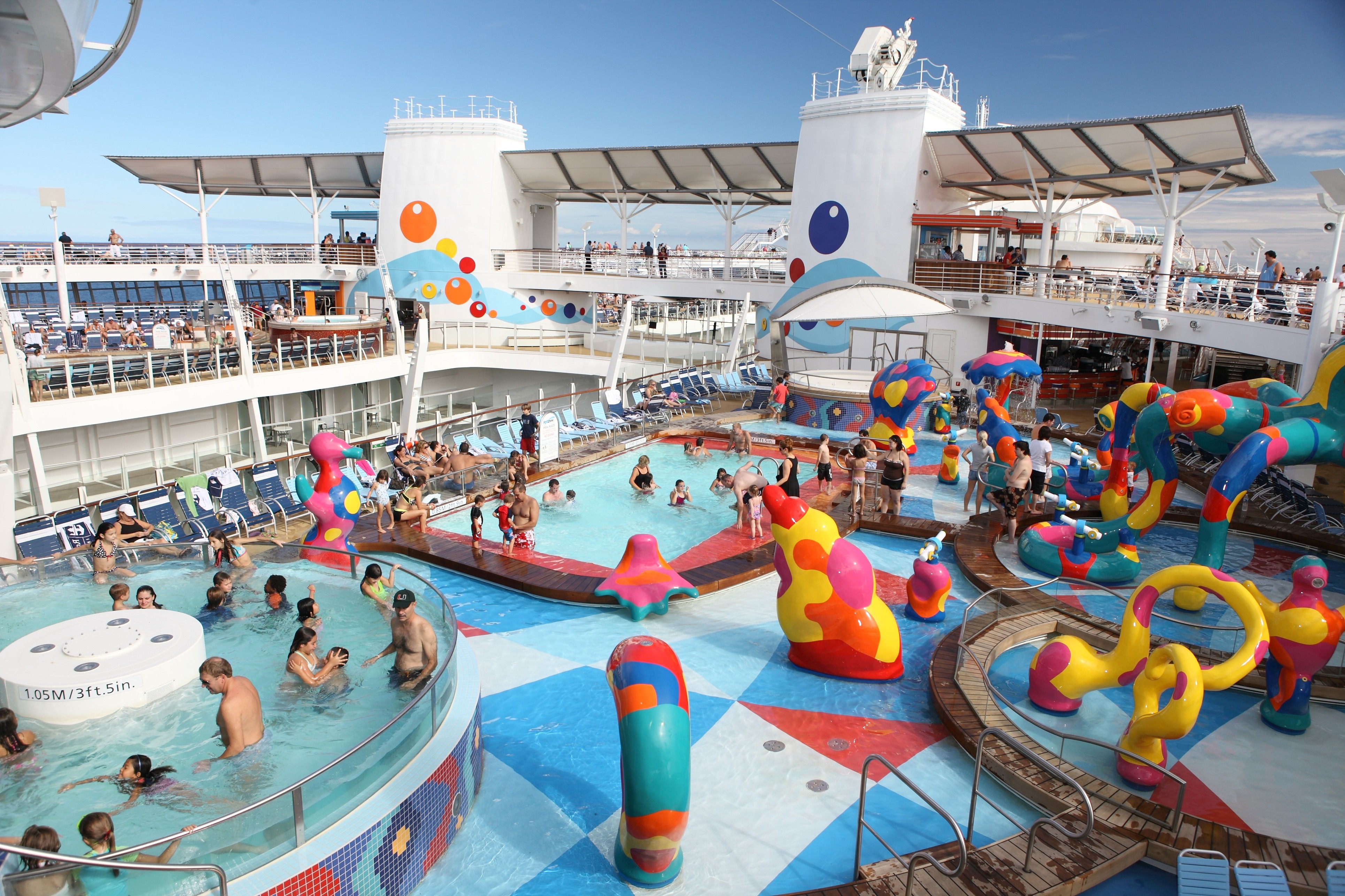 A wide editorial photograph of a dramatic outdoor aqua theater performance on a cruise ship at night, with acrobatic divers mid-air above an illuminated pool, surrounded by a packed amphitheatre audie