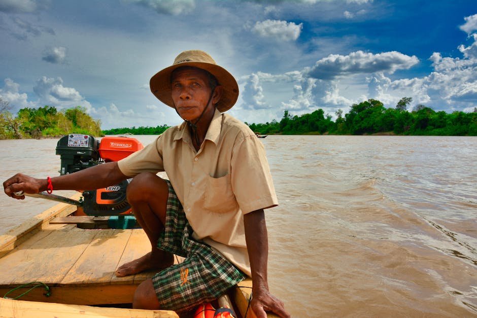 A Cambodian fisherman in a hat operates a wooden boat on the Mekong River under a vibrant cloudy sky.