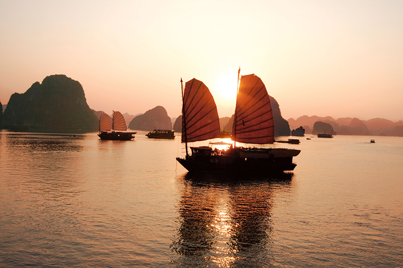 Halong Bay's iconic limestone karsts framing traditional junk boats on calm emerald waters at sunset.