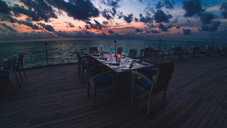 Al fresco oceanfront dining table set against the open sea at sunset, evoking the Waterfront promenade experience aboard Norwegian Breakaway with ambient lighting and ocean views beyond the railing.