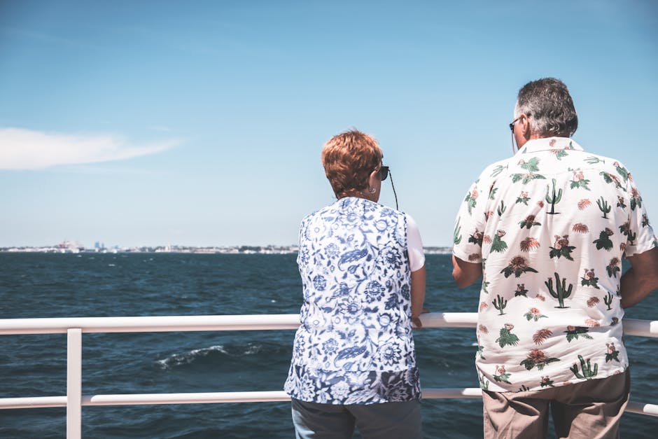 A mature couple seen from behind in casual clothes and sunglasses gazing out at the open sea from a boat deck on a sunny day, capturing the unhurried ease of a long ocean crossing.