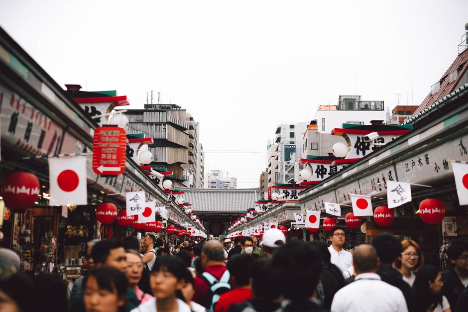 A bustling Japanese street market adorned with traditional flags and paper lanterns, with a diverse crowd of travellers exploring the vibrant local stalls — capturing the culturally immersive port-day experience ideal for Asia cruise passengers.