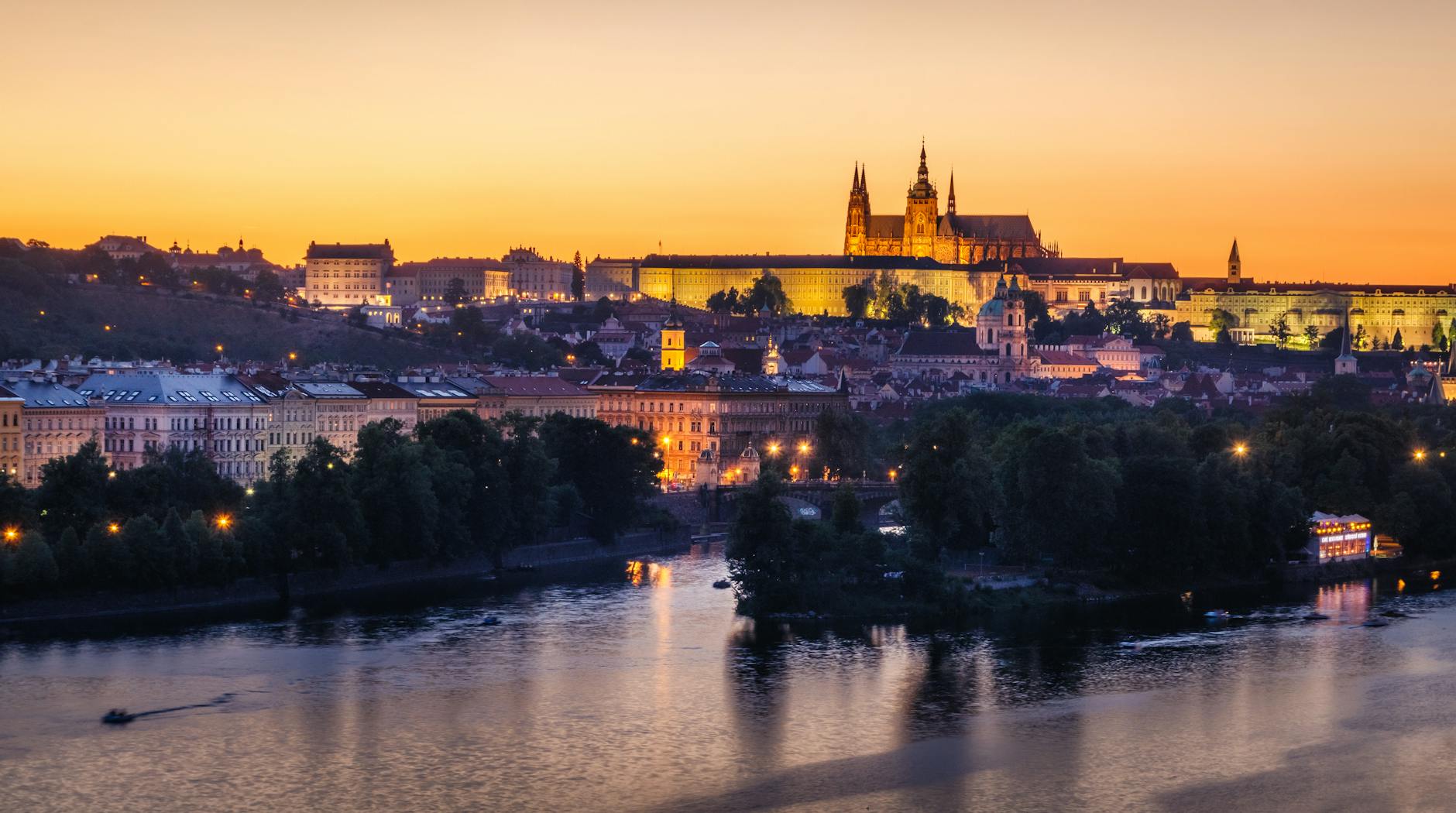 Prague Castle glowing in warm golden light at sunset with the Vltava River flowing in the foreground, evoking the scenic finale of an Elbe River cruise into the Czech capital.