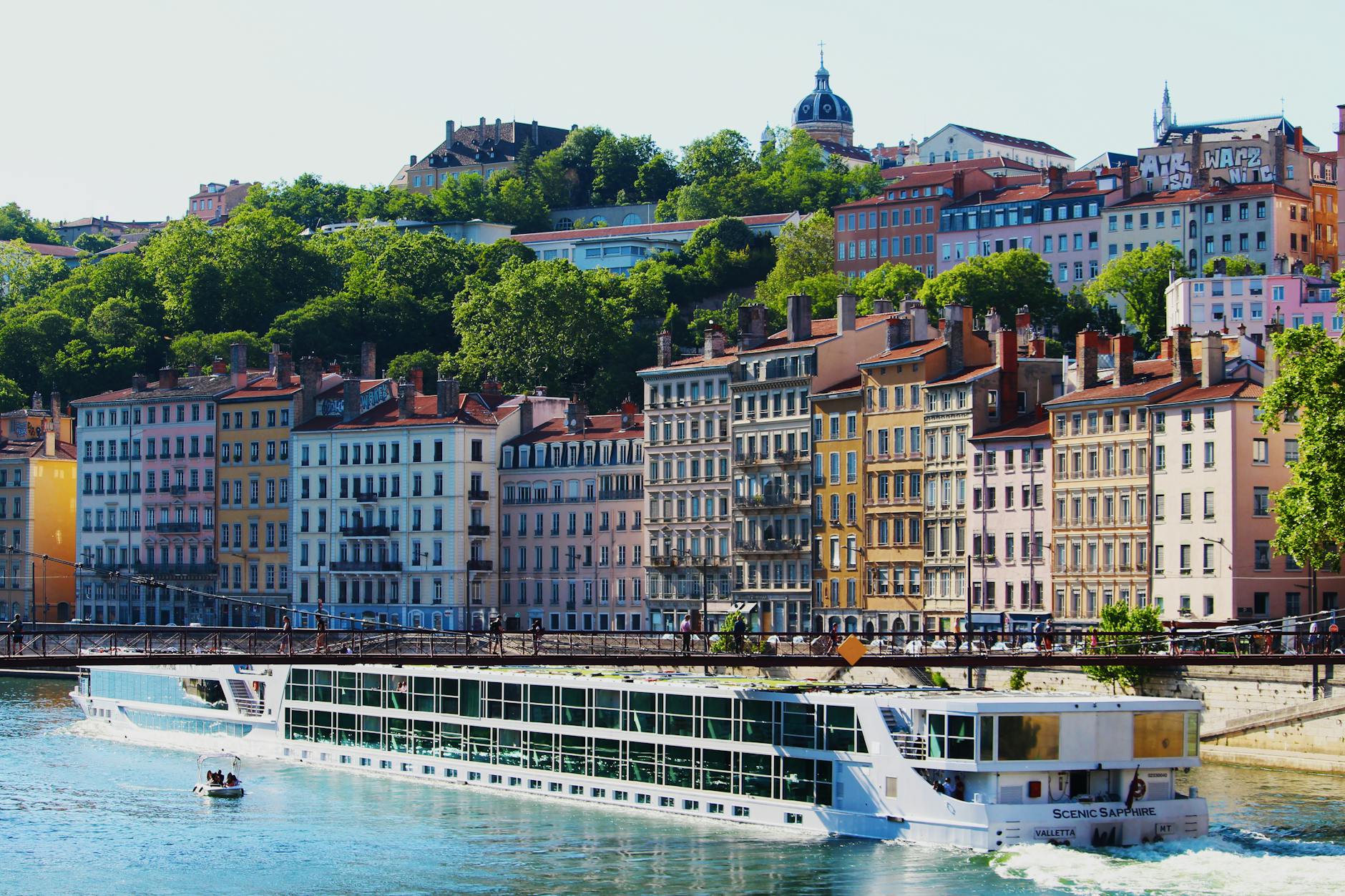 Colorful buildings along the riverbank in bustling Lyon, France, with a scenic cruise ship passing by.