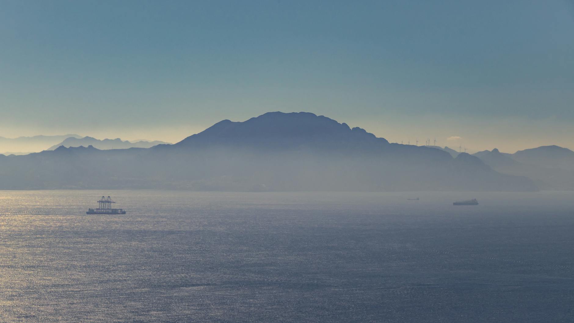 The Strait of Gibraltar seen from the water, with distant mountains along the African and European coastlines and ships visible on the horizon, illustrating the narrow sea passage that repositioning cruises navigate between the Mediterranean and Atlantic.