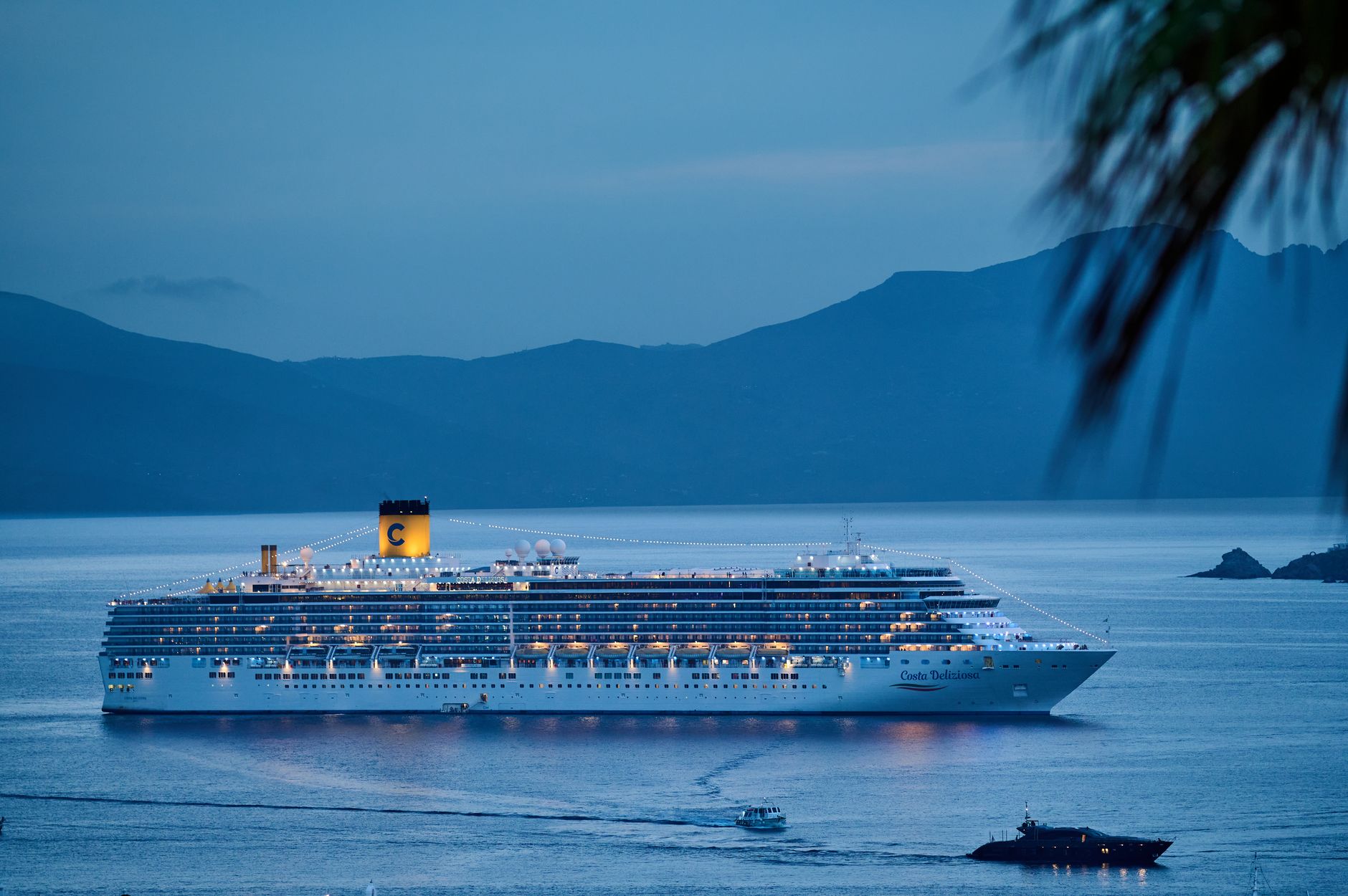 An illuminated cruise ship sailing across calm waters at dusk with mountains framing the harbour, illustrating the variety of cruise lines that depart from this port.