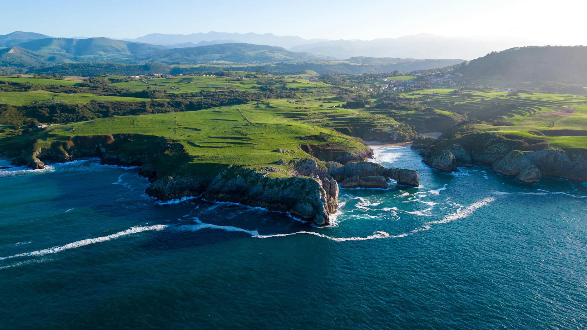Aerial view of the Asturias coastline in northern Spain, where lush green hillsides meet dramatic cliffs and vivid blue Atlantic waves along the Cantabrian coast.