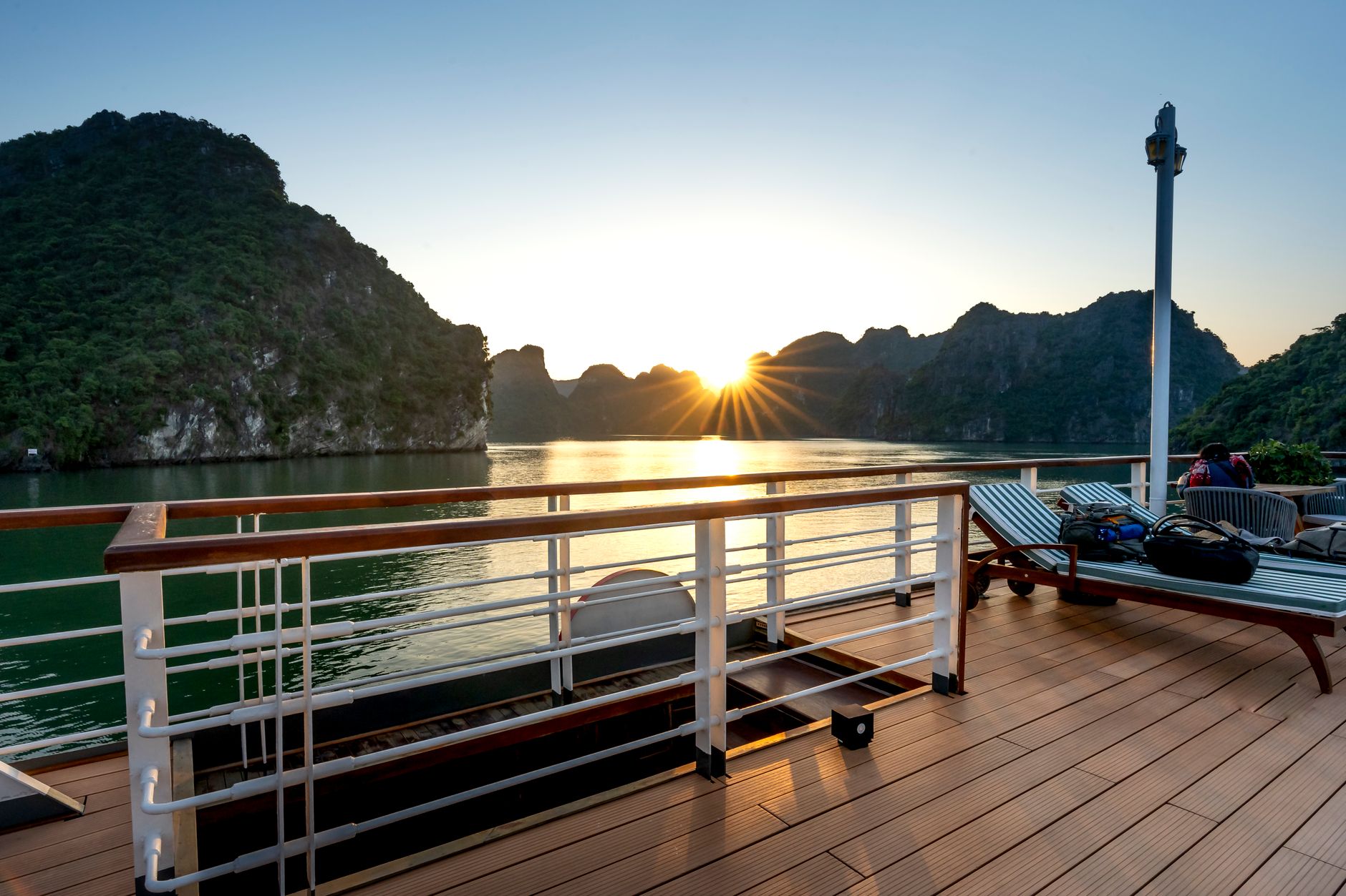A tranquil golden-hour sunset viewed from a river cruise ship's open terrace, with calm water and distant mountains evoking the relaxed, scenic pace of a Rhine wine-country sailing.