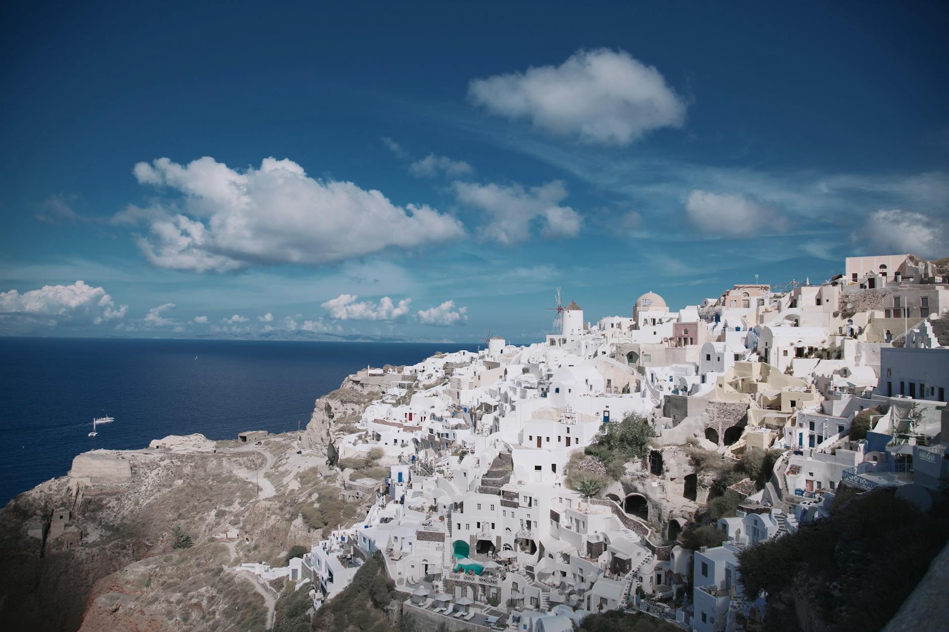 Panoramic golden-hour vista of Oia, Santorini, with whitewashed Cycladic buildings cascading down the caldera cliffside toward the deep blue Aegean Sea under a vibrant sky