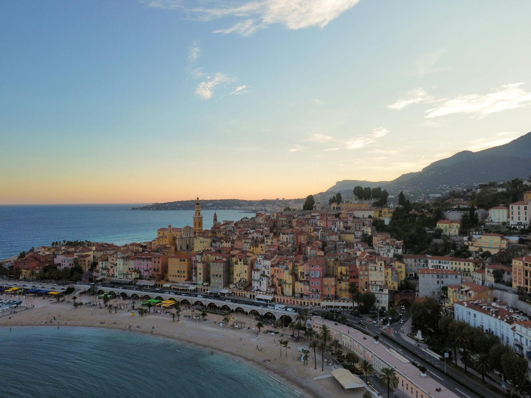 Aerial view of Menton's terracotta-roofed hillside buildings cascading toward the French Riviera coastline, bathed in warm sunset light over the deep blue Mediterranean Sea.