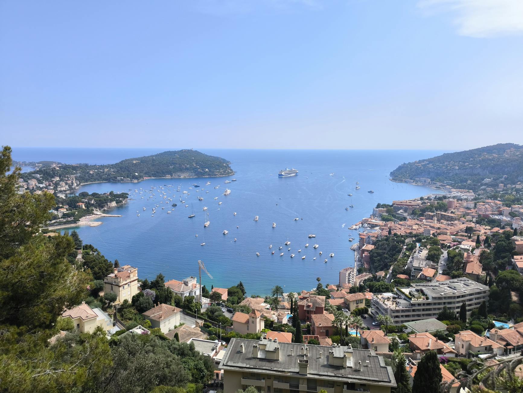 Aerial view of Villefranche-sur-Mer's turquoise bay with colorful pastel buildings cascading down the hillside and a cruise ship anchored among smaller boats in the harbor