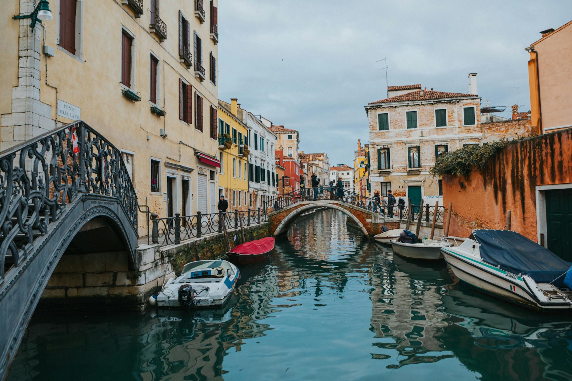 A quiet Venetian canal lined with colorful buildings, their facades reflecting in the still water beneath a stone bridge — the kind of intimate neighbourhood scene that rewards travellers who venture beyond the tourist crowds.