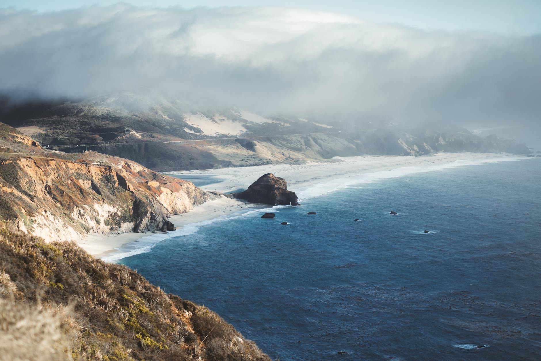 The dramatic Pacific coastline at Big Sur, where rugged cliffs meet the open ocean under sweeping clouds — a defining vista along the southbound coastal cruise route.