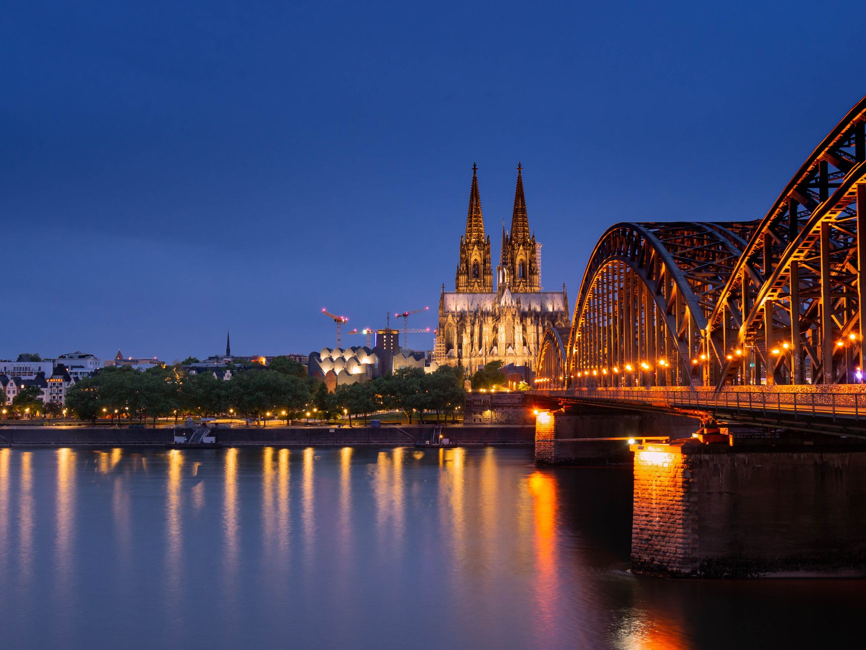 Cologne Cathedral and Hohenzollern Bridge illuminated at night, their lights reflecting across the Rhine River at blue hour