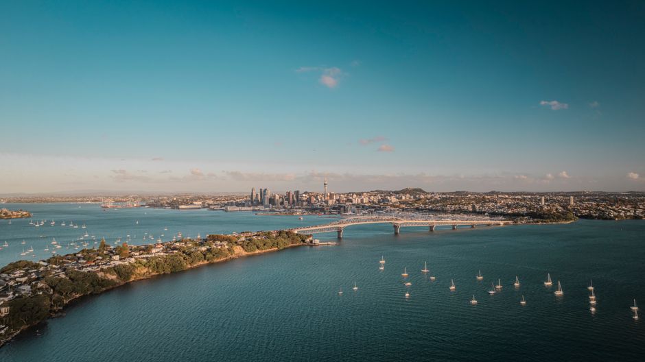 Aerial view of Auckland's skyline and Harbor Bridge stretching over calm harbour waters, establishing the city's waterfront setting for arriving cruise ships.