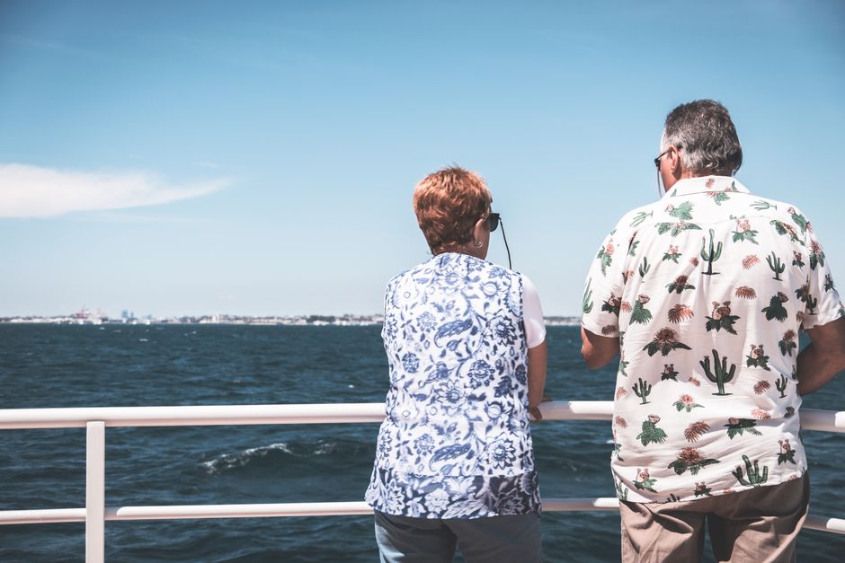 A mature couple seen from behind in casual clothes and sunglasses gazing out at the open sea from a boat deck on a sunny day, capturing the unhurried ease of a long ocean crossing.