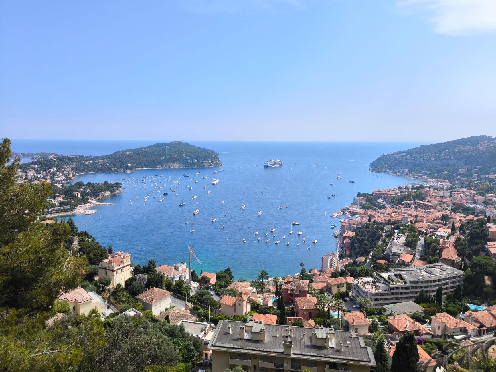 Aerial view of Villefranche-sur-Mer's turquoise bay with colorful pastel buildings cascading down the hillside and a cruise ship anchored among smaller boats in the harbor