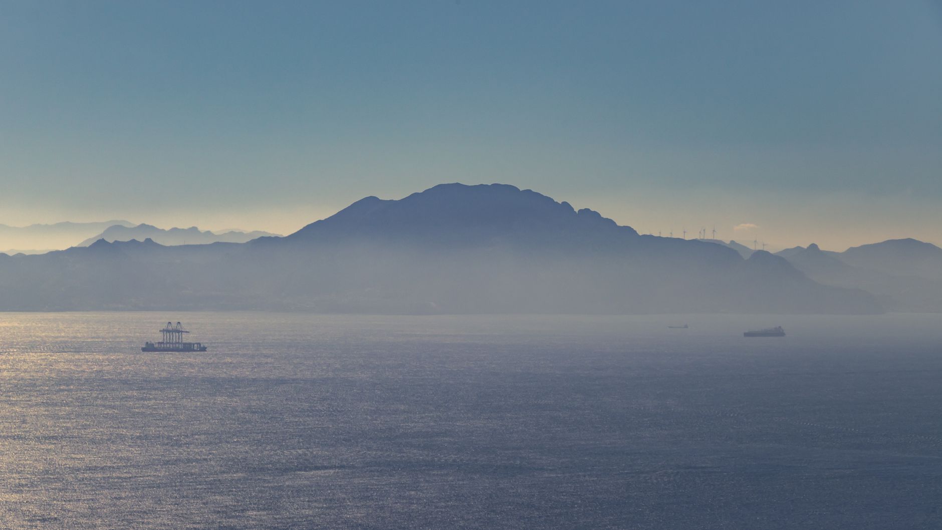 The Strait of Gibraltar seen from the water, with distant mountains along the African and European coastlines and ships visible on the horizon, illustrating the narrow sea passage that repositioning cruises navigate between the Mediterranean and Atlantic.