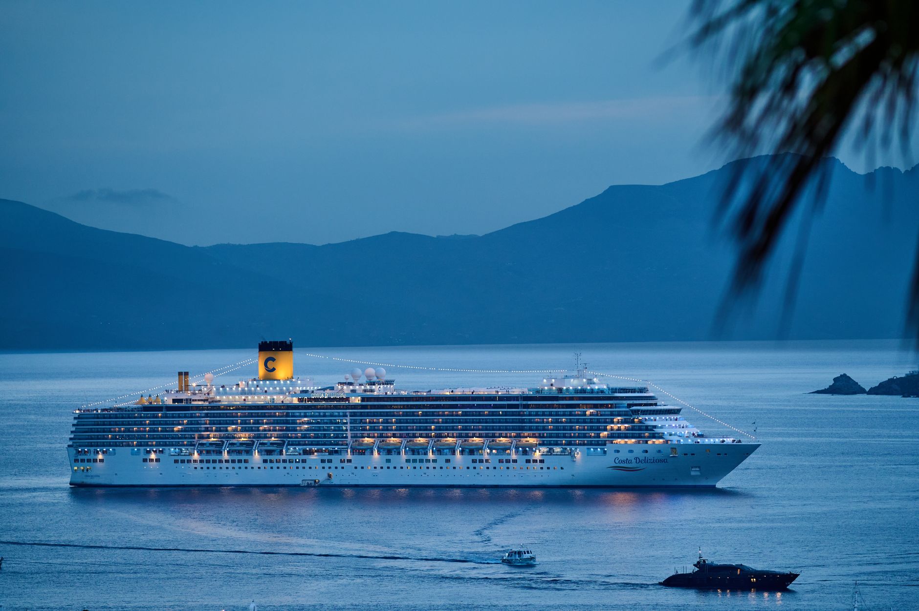 An illuminated cruise ship sailing across calm waters at dusk with mountains framing the harbour, illustrating the variety of cruise lines that depart from this port.
