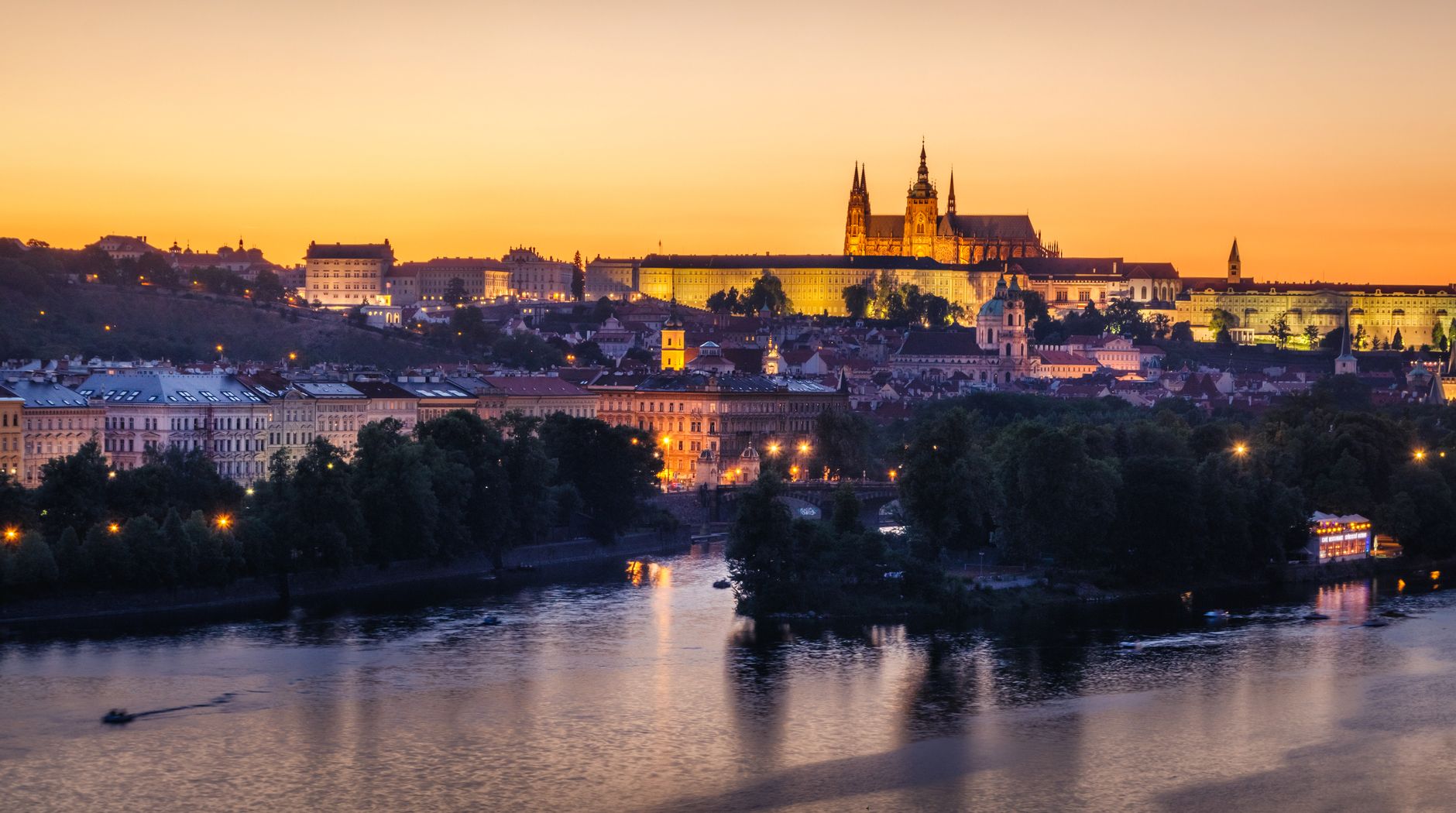 Prague Castle glowing in warm golden light at sunset with the Vltava River flowing in the foreground, evoking the scenic finale of an Elbe River cruise into the Czech capital.
