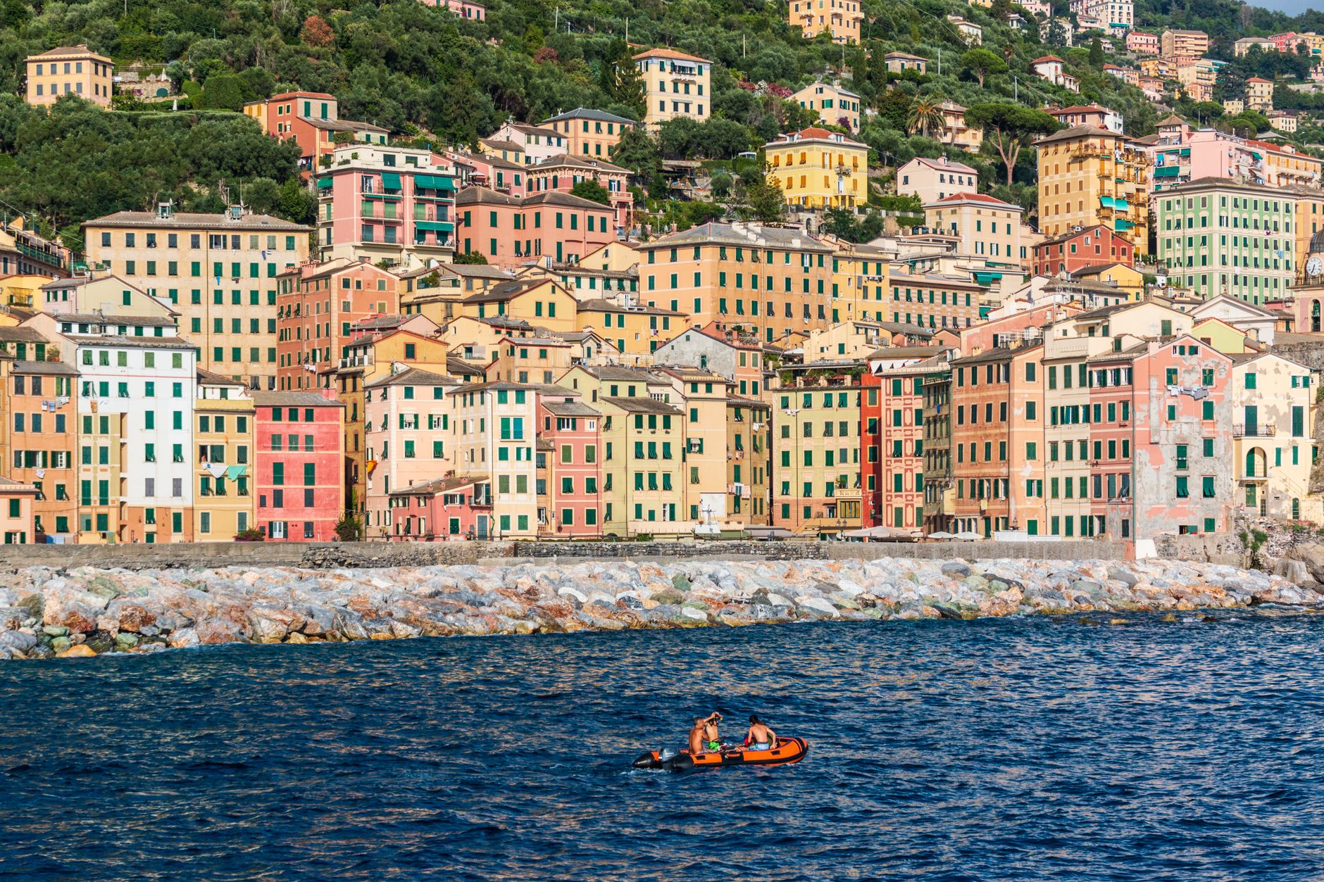 Colourful waterfront buildings line the harbour in Camogli, Liguria, as rowers navigate the blue Mediterranean waters — a quintessential Italian Riviera port town that cruise itineraries frequently visit.