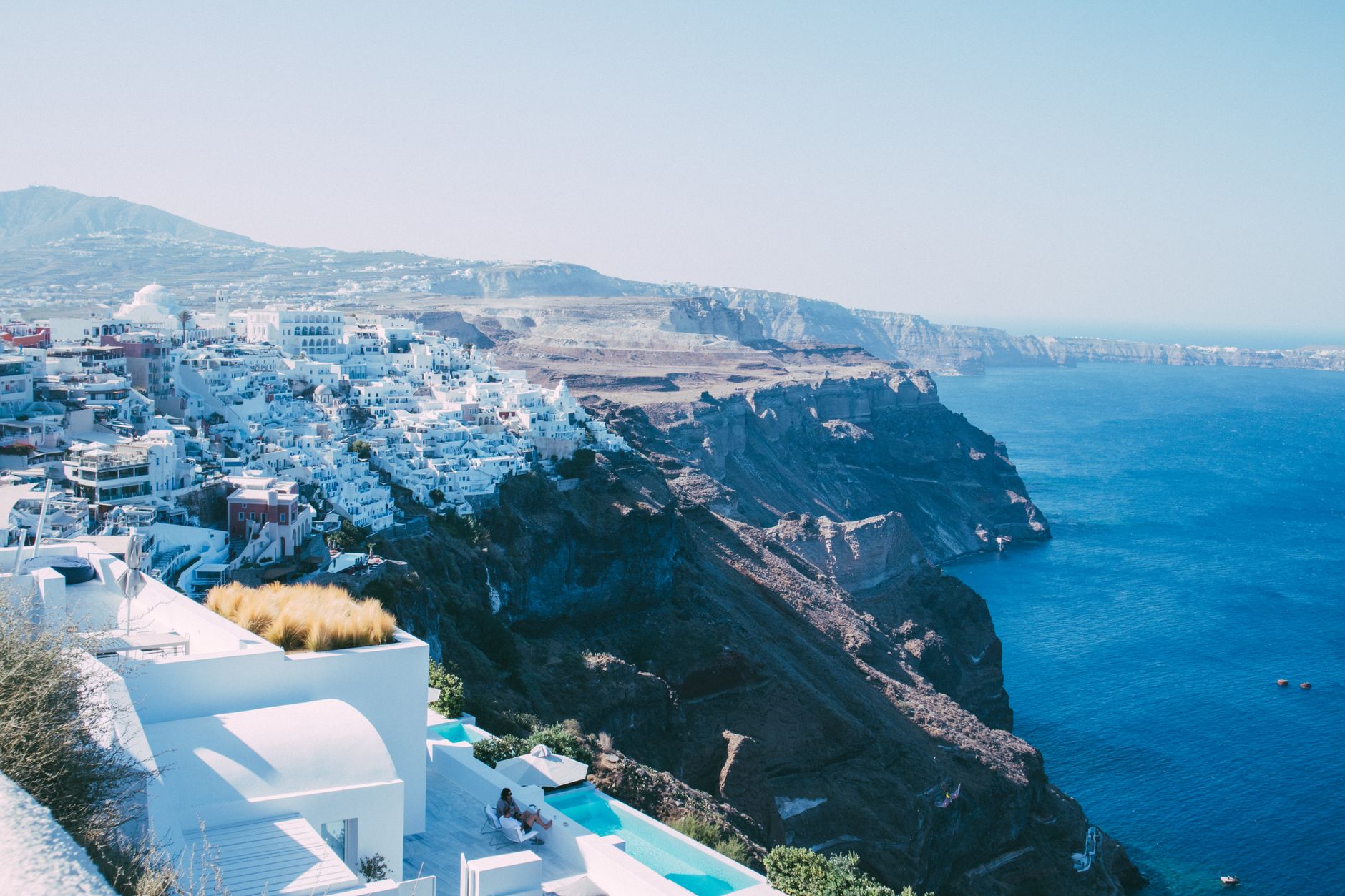 Santorini's iconic white-washed buildings and blue-domed churches cascading down sunlit cliffs above the deep blue Aegean Sea, capturing the essence of Eastern Mediterranean Greek island architecture.
