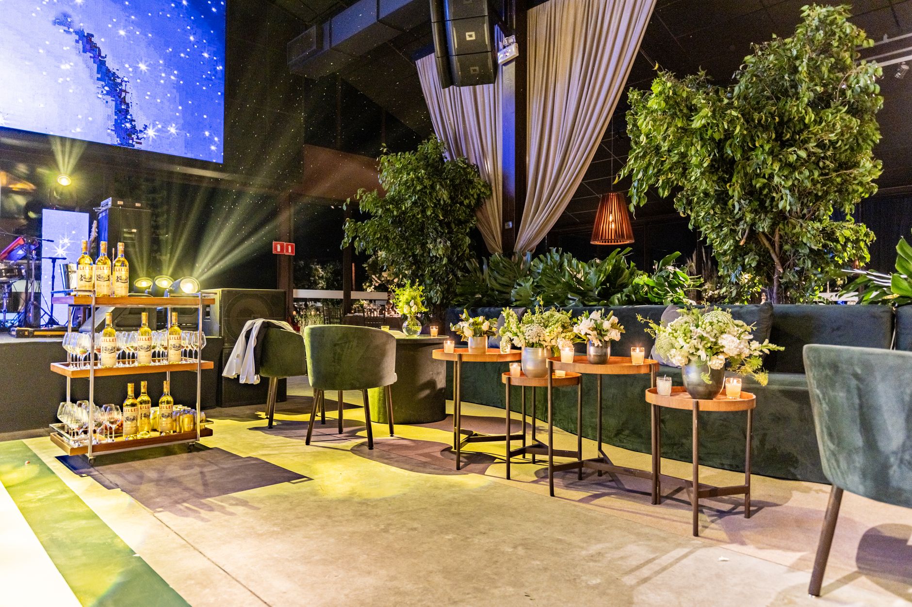 A sophisticated lounge-style interior space with lush greenery, a bar cart, and inviting seating arrangements, evoking the boutique-hotel ambiance found aboard Celebrity Edge-class cruise ships.