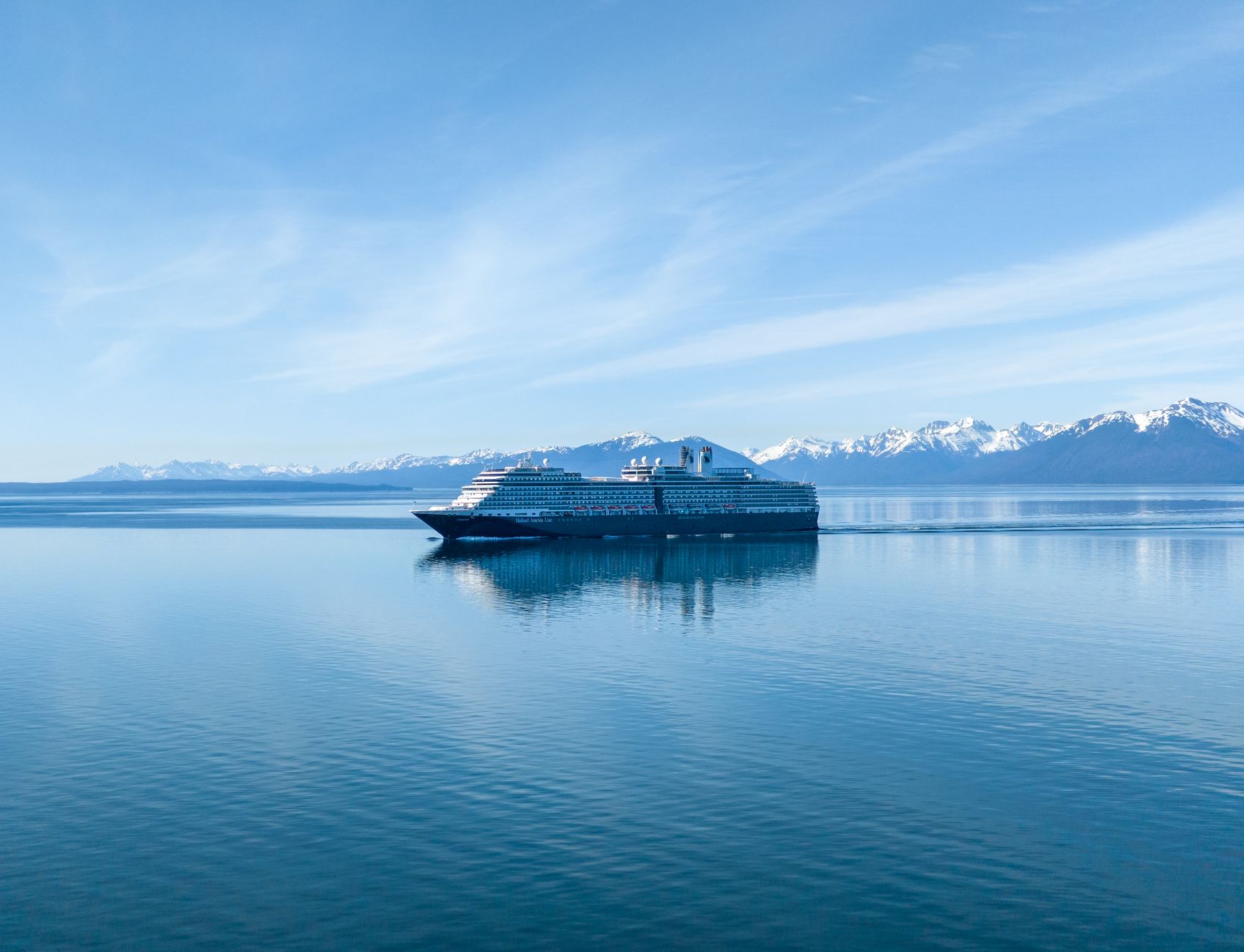 A cruise ship navigating the calm waters of Alaska's Inside Passage near Hoonah, with snow-capped mountains rising behind — a typical scene along the Seward routing operated by ships like Ovation of the Seas.