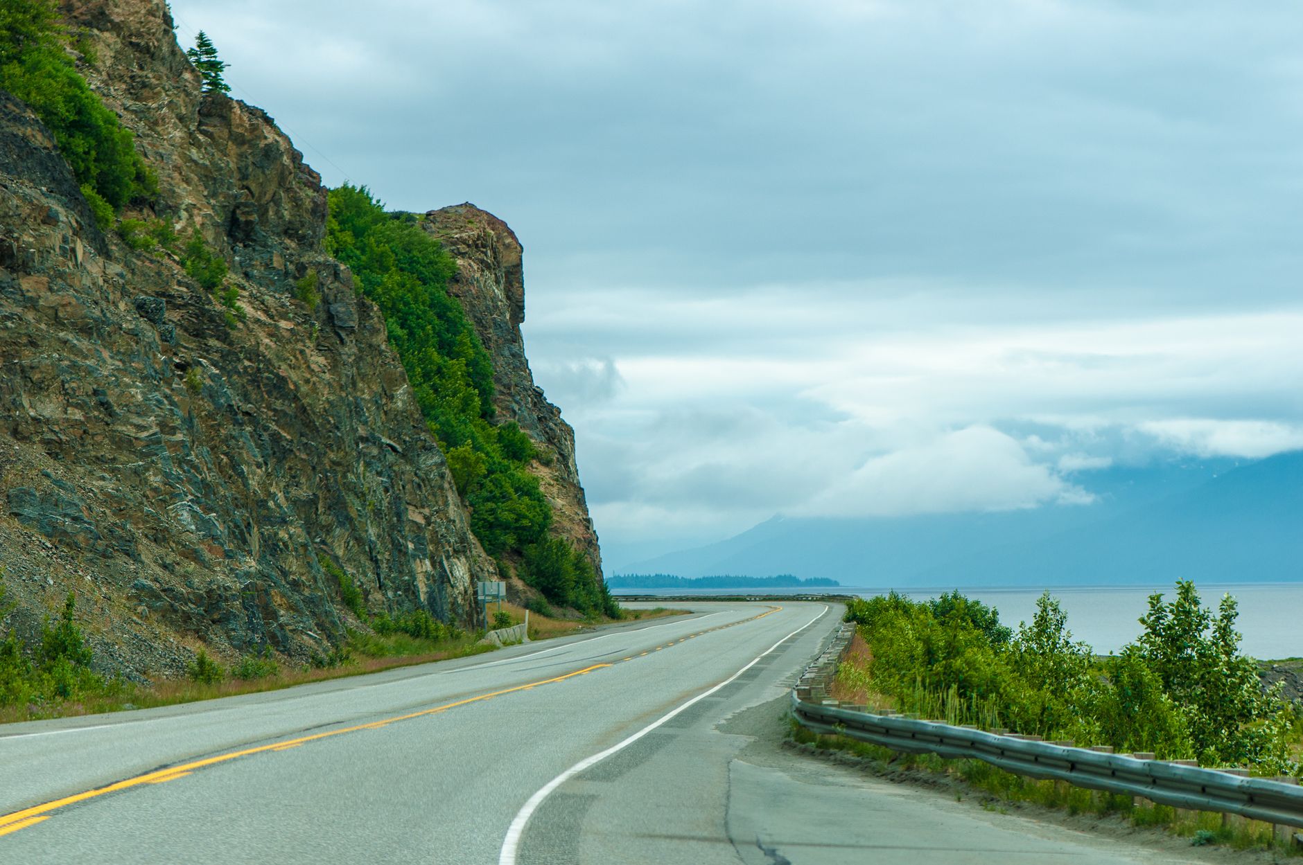 A scenic highway hugging the shoreline beneath towering rock cliffs, evoking the dramatic drive along Turnagain Arm from Anchorage to Seward where mountains rise steeply above calm tidal waters.