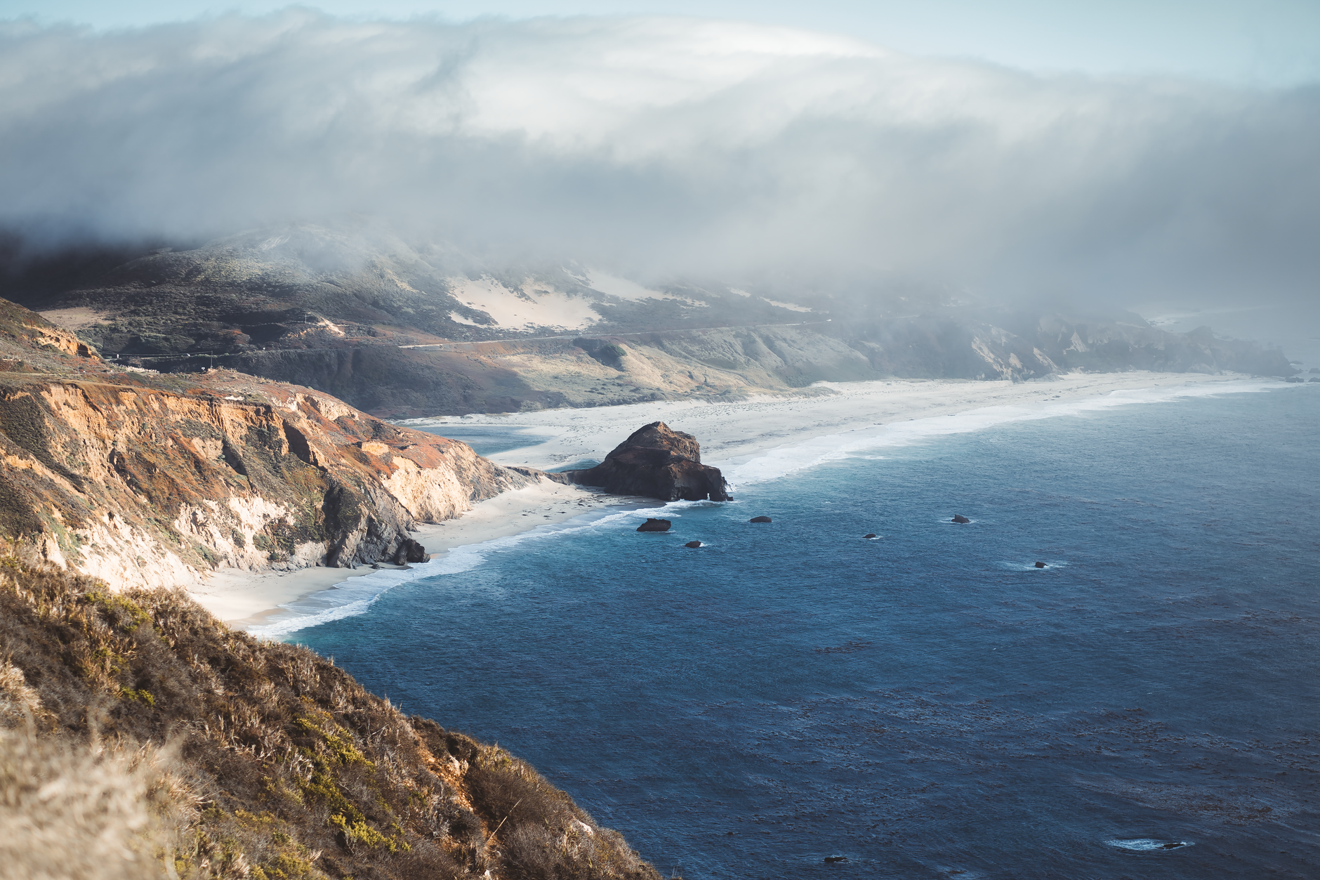 The dramatic Pacific coastline at Big Sur, where rugged cliffs meet the open ocean under sweeping clouds — a defining vista along the southbound coastal cruise route.