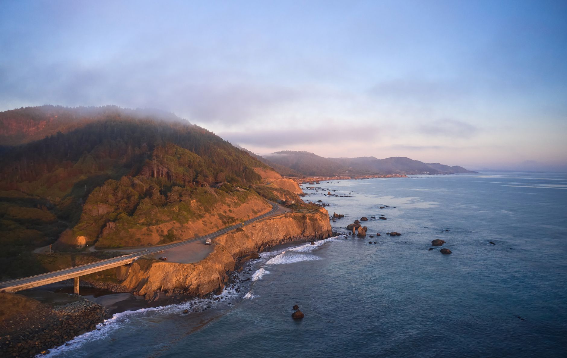 Aerial view of the rugged Pacific coastline at sunset near Westport, evoking the southbound cruise route along forested cliffs and open ocean under golden evening light.
