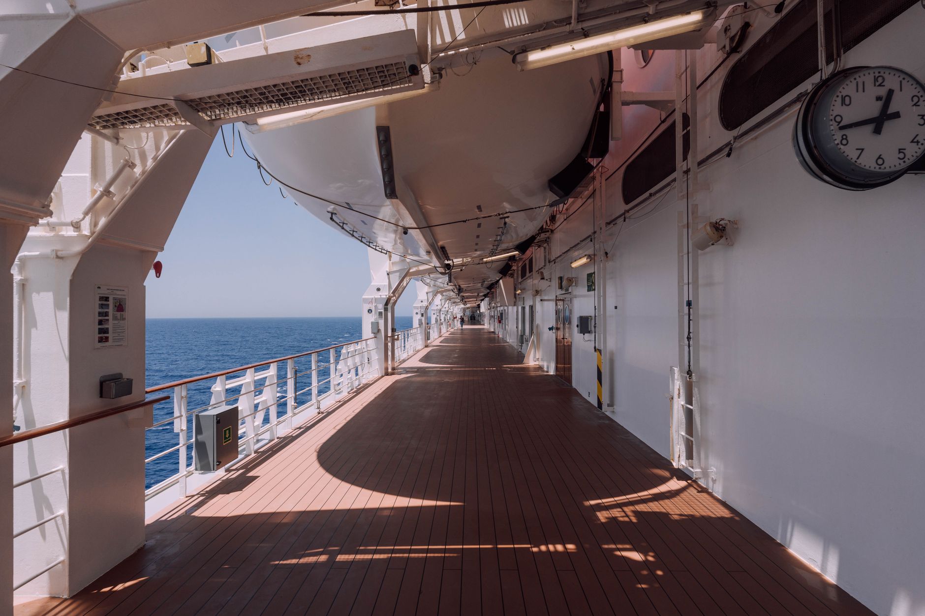 An empty cruise ship promenade deck stretching into the distance, with the open ocean visible beyond the railing and long shadows cast across the deck — evoking the quiet, unhurried atmosphere of a sea day.
