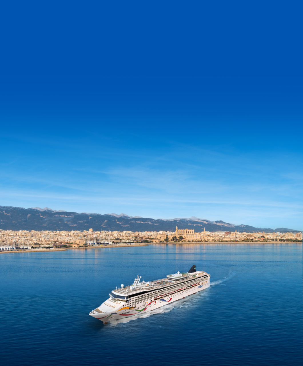 Norwegian Dawn cruise ship anchored in Palma de Mallorca with Mediterranean coastal town and cathedral visible in background under bright sunlight.
