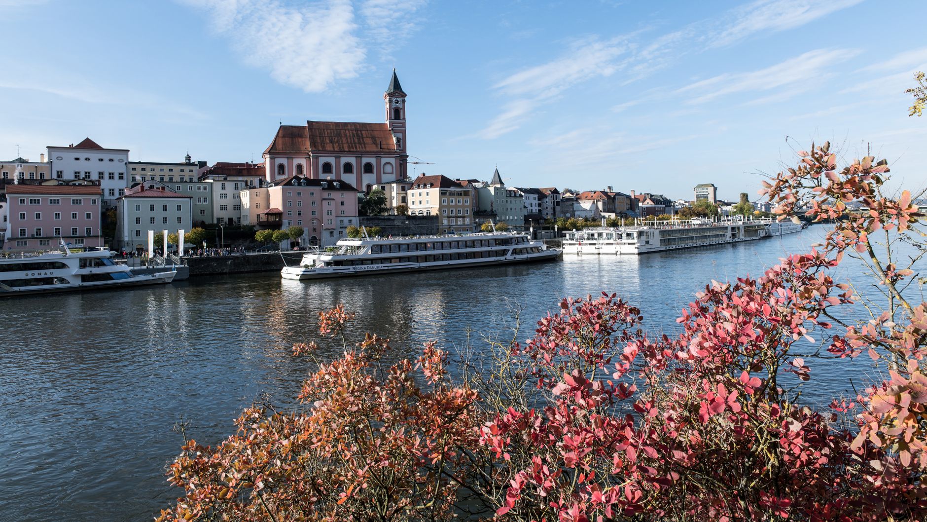 Panoramic view of Passau's historic riverside with docked boats and autumn foliage.